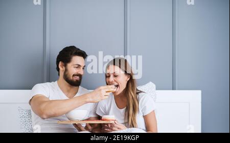 Homme joyeux barbu donnant une belle jeune fille un peu d'un petit déjeuner au lit. Banque D'Images