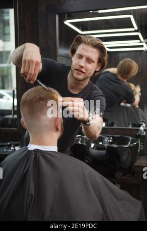 Plan vertical d'un coiffeur joyeux et élégant qui coupe les cheveux d'un client masculin, en appréciant de travailler dans son barbershop Banque D'Images