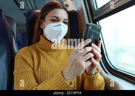 Femme détendue avec le masque KN95 FFP2 en utilisant l'application pour smartphone. Train passager avec masque de protection assis en classe affaires avec envoi de SMS sur mobi Banque D'Images