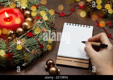 La main d'une femme tient un stylo et écrit dans un bloc-notes. Mise en page. Table en bois du nouvel an avec couronne de sapin, bougies en feu et boules de Noël. Maquette, à Banque D'Images