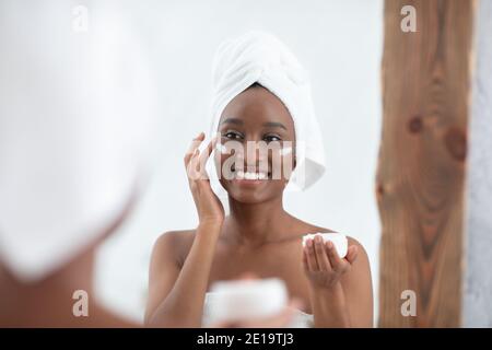 Une adorable femme afro-américaine souriante et mignonne avec une serviette sur la tête après la douche, appliquer de la crème sur le visage Banque D'Images