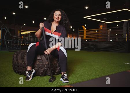 Femme pleine de confiance et de taille souriant à l'appareil photo, assise sur un pneu de roue lourd à la salle de gym, tenant un marteau de traîneau, un espace de copie. Joyeux sport en surpoids Banque D'Images