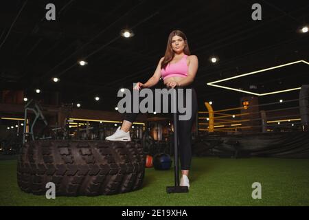 Jeune femme attrayante et de taille plus regardant avec confiance la caméra, se reposant après avoir fait de l'entraînement avec un marteau en traîneau et un énorme pneu de roue lourd. Gym, corps Banque D'Images
