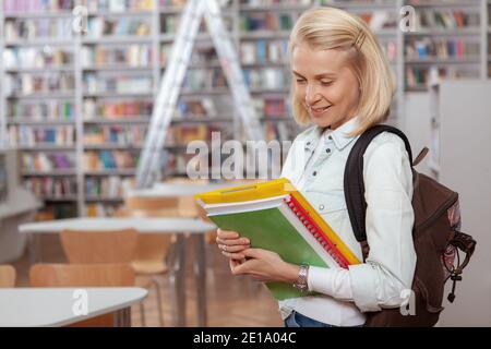 Charmante jeune femme blonde aux cheveux avec sac à dos tenant ses livres à la bibliothèque, espace de copie. Bonne étudiante à la bibliothèque de l'université ou à la librairie Banque D'Images