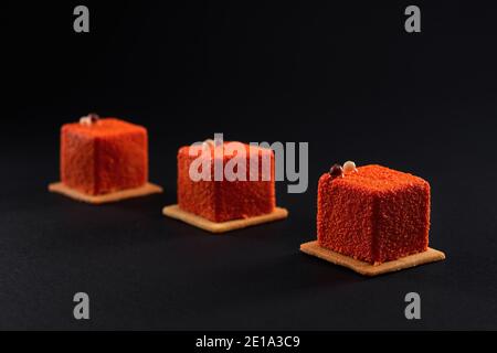 Vue rapprochée d'un dessert orange mat décoré de boules de chocolat isolées sur fond noir de studio. Trois petits gâteaux carrés matte de suite sur des biscuits dans le restaurant. Concept de confiserie. Banque D'Images