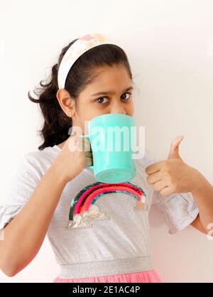 Portrait d'une adorable fille indienne buvant une tasse de vert bleuâtre et montrant les pouces vers le haut. Banque D'Images