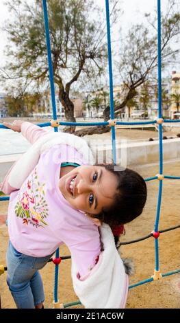 BEAU PORTRAIT D'UNE FILLE SOURIANTE JOUANT HEUREUSEMENT DANS LE GRIMPEUR EN CORDE DANS UN PARC EXTÉRIEUR PENDANT LE COUCHER DU SOLEIL Banque D'Images