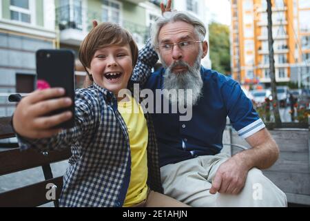 Joyeux grand-père et jeune petit-fils riant, prenant des selfies sur le téléphone samrt dehors dans la ville Banque D'Images