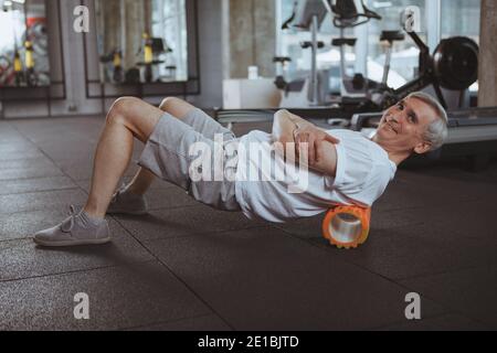 Joyeux homme âgé souriant à l'appareil photo, en utilisant le rouleau en mousse sur son dos. Sportif âgé relaxant ses muscles du dos après avoir fait de l'exercice à la salle de gym. Ma Banque D'Images