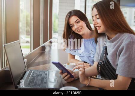 Deux jeunes femmes adorables utilisant un ordinateur portable et un smartphone au café. Des amies travaillant ou étudiant au café du campus Banque D'Images