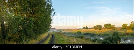 Paysage rural d'été tranquille avec rivière calme, route de campagne et ferme champs le matin Banque D'Images