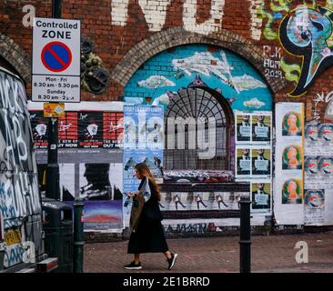 Une femme regardant le téléphone passe devant une fenêtre voûtée grillée dans un mur de briques recouvert de graffiti et d'affiches, Shoreditch, est de Londres, Royaume-Uni. Banque D'Images