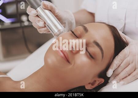 Charmante jeune femme avec une peau douce et soyeuse souriante, recevant un traitement microcourant du visage par un cosmetologiste professionnel. Client féminin qui apprécie le ski Banque D'Images