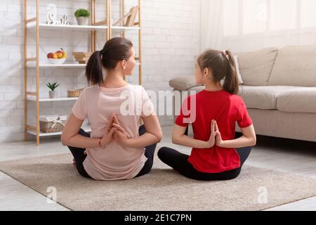 Vue arrière de la jeune mère et de sa fille adolescente faisant du yoga à la maison, assis dans la pose de lotus, faisant un geste de namaste Banque D'Images