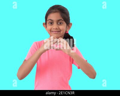 Portrait d'une petite fille indienne mignonne montrant un signe de forme de coeur avec les mains et exprimant l'amour sur fond bleu. Banque D'Images