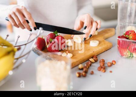 Une jeune femme est coupée sur une planche à découper en bois pour couper de la banane et des fraises fraîches. Banque D'Images