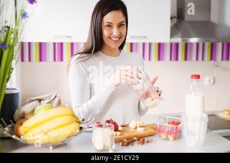 Charmante et charmante jeune femme remplit la tasse du batteur avec des morceaux de fruits frais dans une cuisine très lumineuse. Banque D'Images
