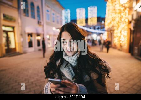 Belle charmante fille en manteau debout dans la rue et regardant le téléphone. Bonne soirée d'hiver. Banque D'Images