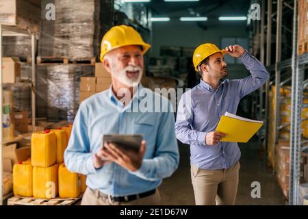 Photo du père et du fils qui marchent dans leur entrepôt avec des casques sur leur tête. Son dans la recherche et la grande étagère pleine de produits et de regarder la focalisation Banque D'Images