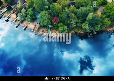 Lac ou rivière avec ciel nuageux bleu de printemps réfléchi et côte de forêt verte avec de petites maisons et des jetées d'eau pour les bateaux, vue aérienne de dessus de drone beau paysage de la nature, copier l'espace Banque D'Images