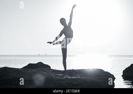 Ballerine jeune en maillot de bain beige danse ballet sur la mer ou la plage de sable de l'océan à la lumière du matin. Concept d'étirement, art, beauté de la nature. Banque D'Images