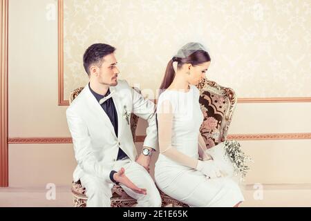 Jeune mariée et marié en colère, homme et femme se sont bouleversés les uns sur les autres assis dans des fauteuils regardant le côté isolé sur fond classique. Conflit entre les deux Banque D'Images