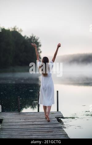 La jeune femme respire profondément en se tenant sur une jetée en bois au lever du soleil le matin au bord d'un lac brumeux Banque D'Images