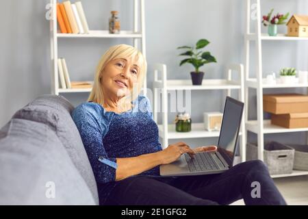 Femme blonde d'âge moyen souriante assise sur un canapé et travaillant en ligne sur pc portable à la maison Banque D'Images