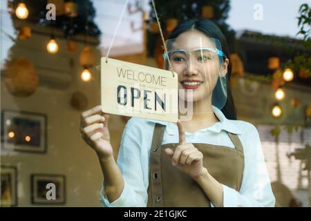 La propriétaire d'entreprise asiatique femme porte un écran facial de protection suspendu panneau ouvert à son restaurant / café, ouvert à nouveau après verrouillage pour un nouveau style de vie normal Banque D'Images