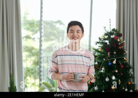 Joyeux jeune homme d'affaires vietnamien debout près du sapin, tenant une tasse de café Banque D'Images