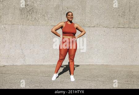 Portrait complet d'une femme en bonne santé debout à l'extérieur. Femme en vêtements de sport debout avec les mains sur les hanches, regardant l'appareil photo et souriant. Banque D'Images