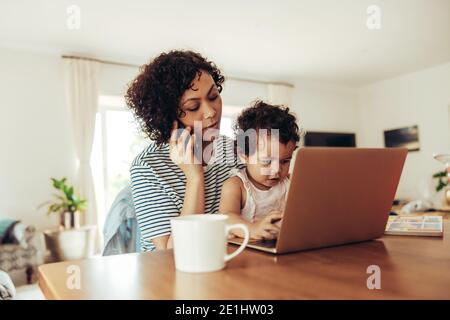 Femme parlant au téléphone avec son bébé assis à une table. La mère qui travaille parle au téléphone portable avec bébé à la maison. Banque D'Images