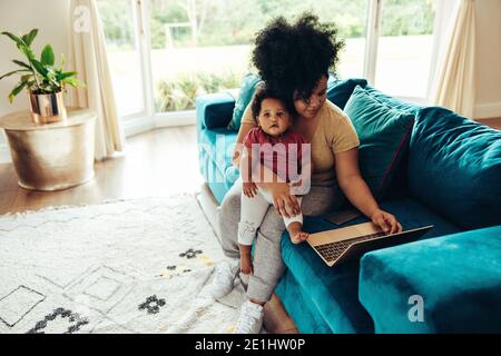 Femme assise avec un bébé sur le canapé et utilisant un ordinateur portable. Mère avec son bébé travaillant à la maison. Banque D'Images