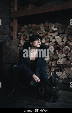 Femme à la mode souriant et se pliant devant un pile de morceaux de bois avec son bouledogue français Banque D'Images