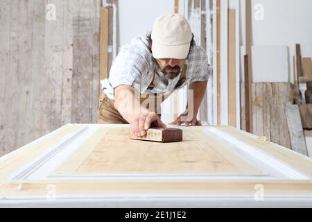 Charpentier mâle travaillant le bois dans l'atelier de menuiserie, sablant une porte en bois avec du papier de verre, portant l'ensemble et le chapeau Banque D'Images