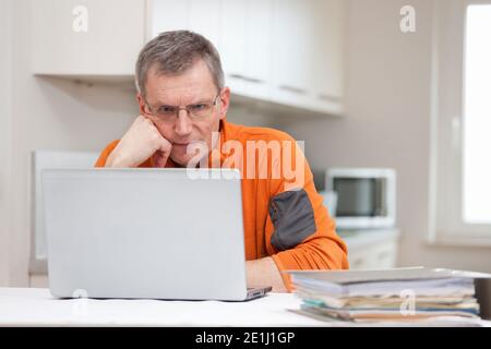 Homme pensif mature travaillant à distance de la maison avec un ordinateur portable et fichiers - en arrière-plan une cuisine lumineuse Banque D'Images