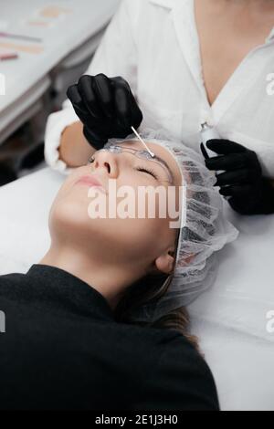 Gros plan du visage de la femme avec les yeux fermés. Les mains du maître en gants noirs appliquent de la crème anesthésique aux sourcils avant le mak permanent Banque D'Images