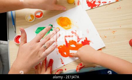 Vue de dessus d'un petit garçon avec dessin de mère et peinture avec les mains couvertes de peinture colorée sur papier blanc. Concept de l'éducation de l'enfant, de la créativité Banque D'Images