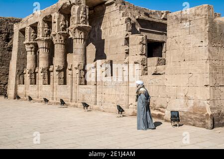 Edfu. Un homme local en robe arabe traditionnelle attend les touristes en tant que guide non officiel ou pose pour des photos au Temple 200BC d'Horus le Dieu faucon, situé à mi-chemin entre Louxor et Assouan sur le Nil et la ville d'Edfu célèbre pour ses énormes pylônes, colonnes finement décorées, hiéroglyphes cartouche racontant l'histoire de panneaux muraux et œuvres d'art fines sur ses plafonds. Le temple a eu la chance que la majorité de celui-ci ait été couvert de sable soufflé par le vent préservant ainsi le temple des éléments pendant des siècles. Banque D'Images