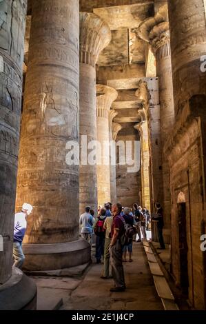 Edfu. Les touristes contemplez quelques-unes des puissantes colonnes finement sculptées dans le hall Hypostyle au temple de Horus le Dieu Falcon, situé à mi-chemin entre Louxor et Assouan sur le Nil et la ville d'Edfu célèbre pour ses énormes pylônes, colonnes finement décorées, cartouche hiéroglyphes histoire racontant des panneaux muraux, et des œuvres d'art sur ses plafonds. Le temple a eu la chance que la majorité de celui-ci ait été couvert de sable soufflé par le vent préservant ainsi le temple des éléments pendant des siècles. Banque D'Images