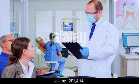 Médecin prenant des notes sur le presse-papiers au sujet des problèmes dentaires des patients assis sur une chaise dans la salle d'attente de la clinique stomatologique. Assistant préparant la vieille femme à l'examen en arrière-plan Banque D'Images