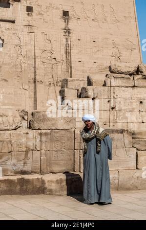 Edfu. Un homme local en robe arabe traditionnelle s'efforce de gagner de l'argent en posant pour les touristes au Temple 200BC d'Horus le Dieu faucon, situé à mi-chemin entre Louxor et Assouan sur le Nil et la ville d'Edfu célèbre pour ses énormes pylônes, finement décorées colonnes, cartouche hiéroglyphes histoire racontant des panneaux muraux, et des œuvres d'art sur ses plafonds. Le temple a eu la chance que la majorité de celui-ci ait été couvert de sable soufflé par le vent préservant ainsi le temple des éléments pendant des siècles. Banque D'Images