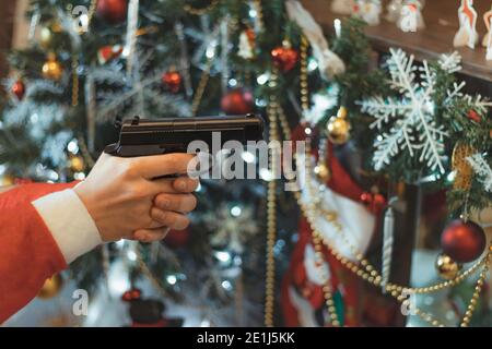 Main en costume le Père Noël tient le fusil sur fond d'arbre de Noël. Main Mans tenant le pistolet chargé. Concept de Joyeux Noël et de la Saint-Sylvestre. Banque D'Images