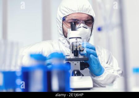 Microbiologiste chercheur portant des epi et des lunettes de protection dans un laboratoire de médecine regardant au microscope. Scientifique en costume de protection assis sur le lieu de travail utilisant la technologie médicale moderne pendant l'épidémie mondiale. Banque D'Images