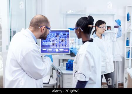 Un scientifique et un médecin africain étudiant la guérison d'un virus dangereux sur ordinateur dans un laboratoire très fréquenté. Une équipe multiethnique de chercheurs médicaux travaillant ensemble dans un laboratoire stérile portant des lunettes et des gants de protection. Banque D'Images