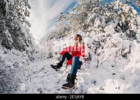 Jeune femme en blouson rouge assise parmi les arbres enneigés dans la forêt d'hiver. Voyageur femme avec des arbres de neige par temps ensoleillé Banque D'Images