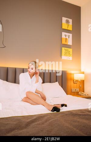 Jolie fille blonde en peignoir blanc est assise sur un lit; elle porte des chaussures. Banque D'Images