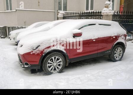 Voiture rouge moderne recouverte de neige blanche garée près de la maison dans la rue en hiver Banque D'Images