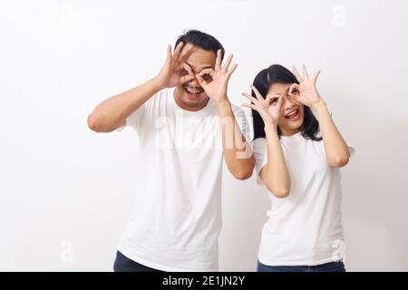 Jeune beau couple asiatique ensemble sur un arrière-plan isolé faisant un geste ok comme des jumelles, les yeux regardant à travers les doigts. Expression folle. Banque D'Images