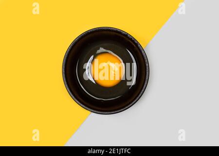 Un œuf cru dans une assiette noire dans un sommet afficher sur fond jaune et gris Banque D'Images